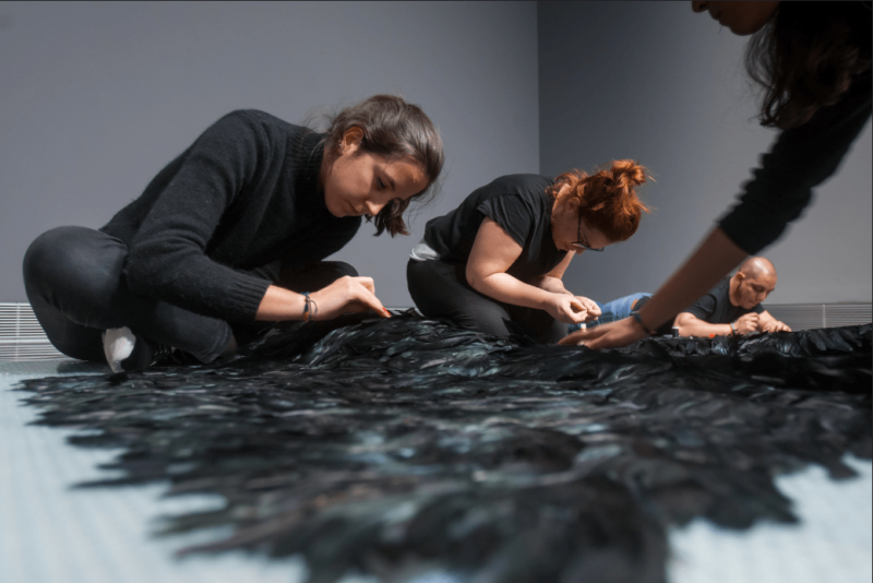 En la imagen varios estudiantes tejen un manto de plumas negras, que forman parte de una pieza de la artista Cecilia Paredes. 