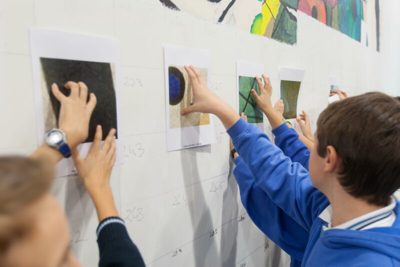 En la imagen los niños sostienen piezas que ellos mismos han confeccionado, y las colocan en el gran mural de pared que se realizará con sus aportaciones. 