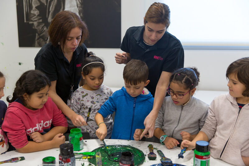 En la imagen, alrededor de una mesa, varios niños y niñas utilizan pinturas y pinceles con el apoyo de las monitoras. Fotografía de @Manuel Castells.