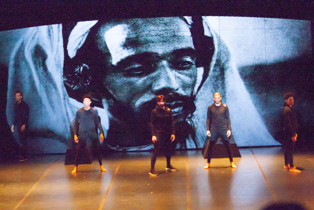 Sobre el escenario, varios bailarines danzan ante una imagen de una fotografía de un africano, forma parte de la coreografía Atlas 2 de Dani Pannullo