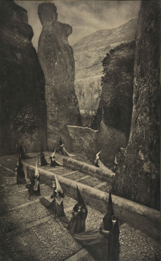 Los penitentes de Viernes Santo embozados con capirotes suben apoyándose sobre el báculo de las velas por las empinadas cuestas en escalera de Cuenca como parte de la procesión de Viernes Santo. Al fondo un impresionante paisaje de roca, muros escarpados propios del recorrido. La imagen presenta los tonos y texturas característicos del autor. 