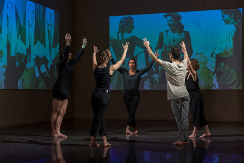 In front of two large screens showing the photos Las Roncalesas by Ortiz Echagüe, several dancers dance in a circle with their arms raised in the air.