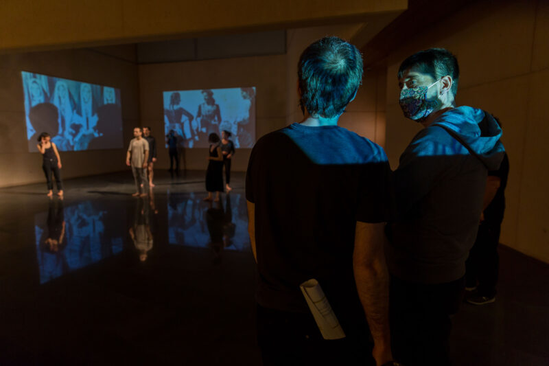 The dancer Jon Maya talks to the director in room -1 of the Museum. On the two walls in the corner of the room are projected two photographs of Las Roncalesas by José Ortiz Echagüe taken in the 1919 flu, in front of which the barefoot dancers are rehearsing.