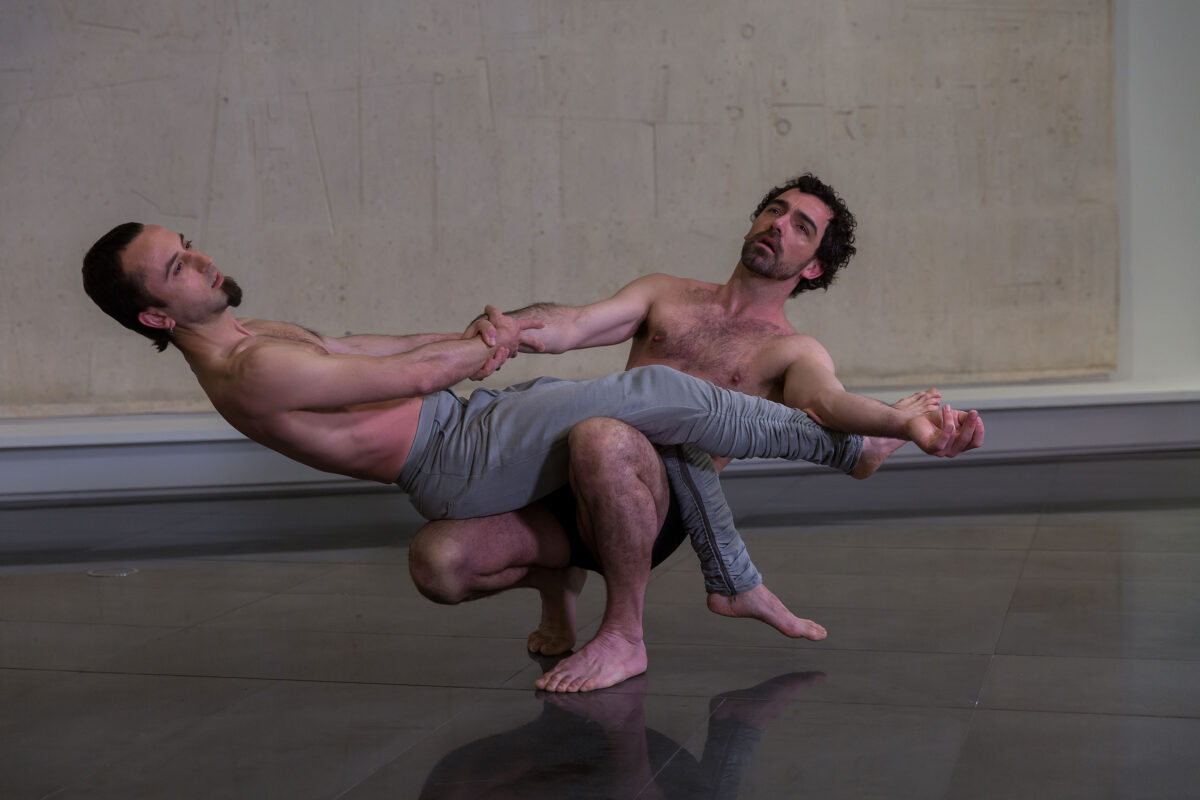 Two dancers in a moment of dance before the piece Homenaje a Bach by Jorge Oteiza. The first dancer squats down and holds a second dancer in balance. Both look upwards in an attitude of prayer.